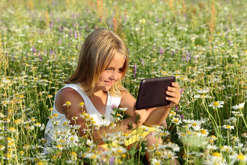 Śliczna uśmiechnięta dziewczynka bawi się tabletem wśród polnych kwiatów. - obrazy, fototapety, plakaty 