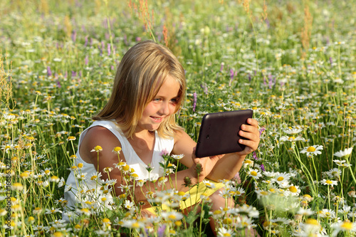 Śliczna uśmiechnięta dziewczynka bawi się tabletem wśród polnych kwiatów.