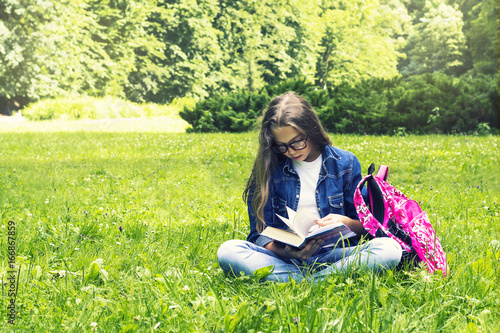 Beautiful blonde schoolgirl girl in jeans shirt reading a book on grass with a backpack in the park © vallerato