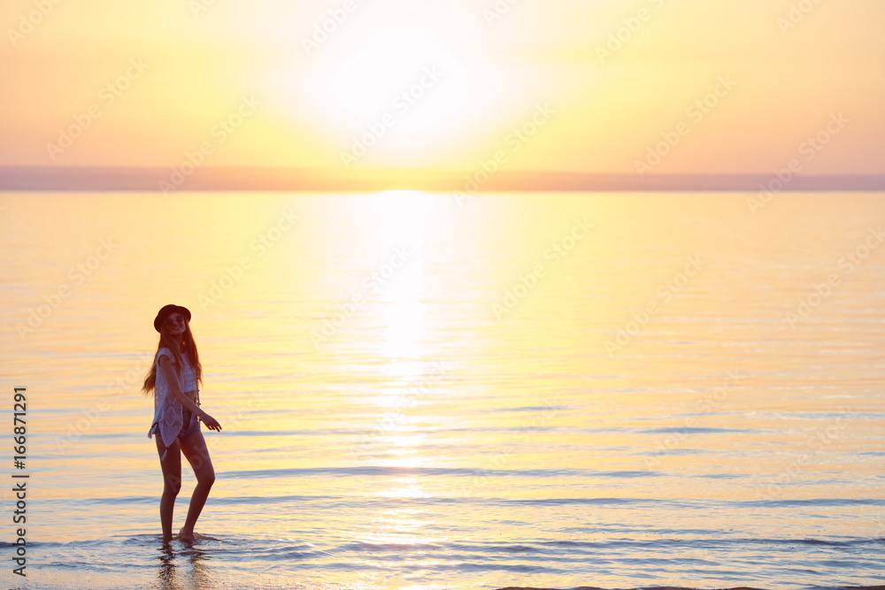 Young, beautiful girl walking at the beach at sunset. Stylish woman with long hair standing in blouse and jeans shorts