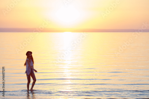 Young  beautiful girl walking at the beach at sunset. Stylish woman with long hair standing in blouse and jeans shorts