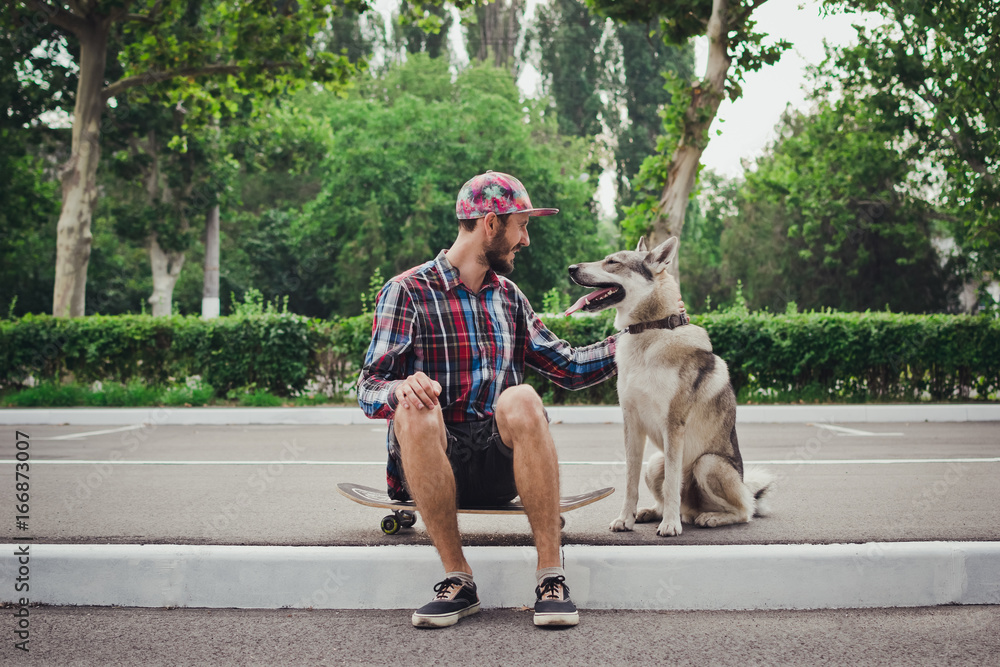 Obraz premium Young hipster man sitting on skateboard with siberian husky dog on street road