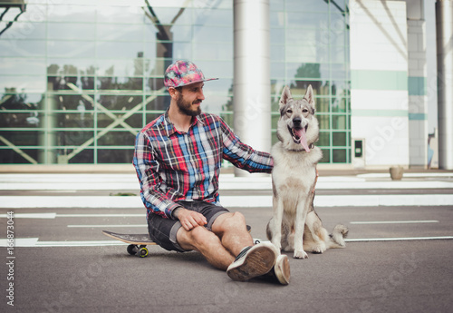 Young hipster man sitting on skateboard with siberian husky dog on street road