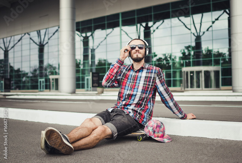 Portrait of young hipster man sitting on the skateboard on street road and listen music