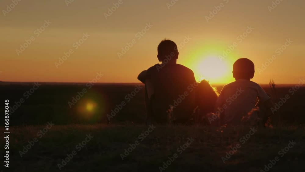 Silhouettes of two rural boys. Sit next to the dog, admiring the beautiful sunset together Silhouettes of two rural boys. Sit next to the dog, admiring the beautiful sunset together
