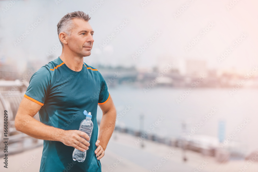 Plakat Portret zdrowego atletycznego w średnim wieku mężczyzna z dysponowaną ciało mienia butelką odświeżająca woda, odpoczywający po treningu lub biegać. na riverside. kolor vintage