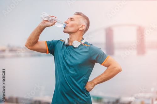 Portrait of healthy athletic middle aged man with fit body holding bottle of refreshing water, resting after workout or running. on the riverside. vintage color