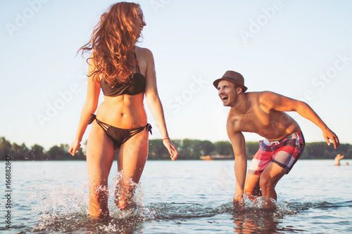 Happy couple runs through waves on sunlit beach, splashing the water in the sea.