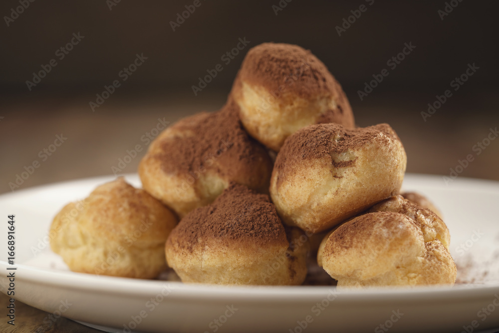 closeup shot of profiteroles covered with cocoa powder on white plate on wooden table closeup shot of profiteroles covered with cocoa powder on white plate on wooden table