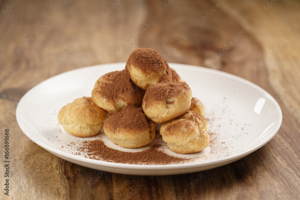 profiteroles covered with cocoa powder on white plate on wooden table profiteroles covered with cocoa powder on white plate on wooden table