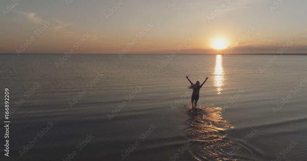 Aerial shot over teen girl barefoot running and raising arms in water on baltic sea beach in sunset time