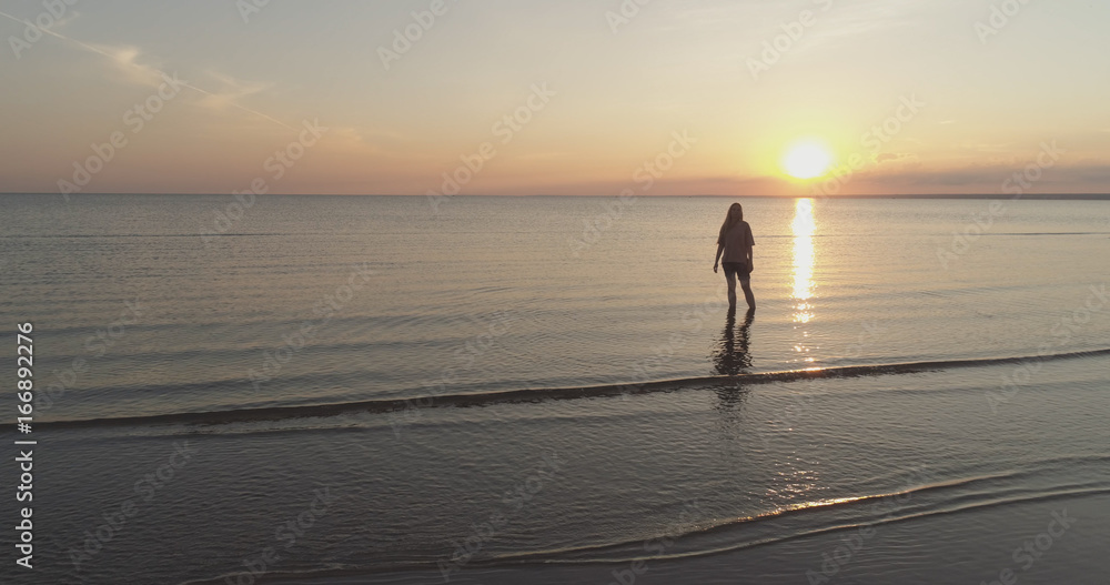 Aerial shot over teen girl barefoot stand in water on baltic sea beach in sunset time