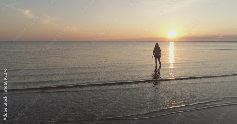 Aerial shot over teen girl barefoot stand in water on baltic sea beach in sunset time Aerial shot over teen girl barefoot stand in water on baltic sea beach in sunset time