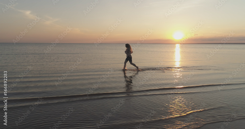 Aerial shot over teen girl barefoot running in water on baltic sea beach in sunset time