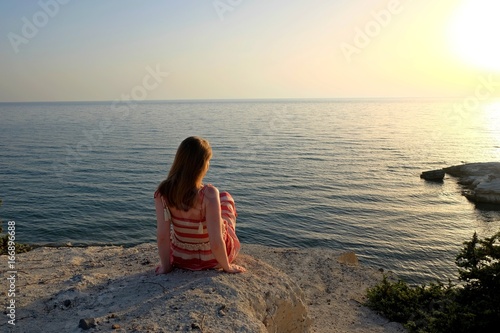 Tranquil seascape scene with girl which looking at the sun sitting on the rock at the beach. Kos island Greece.