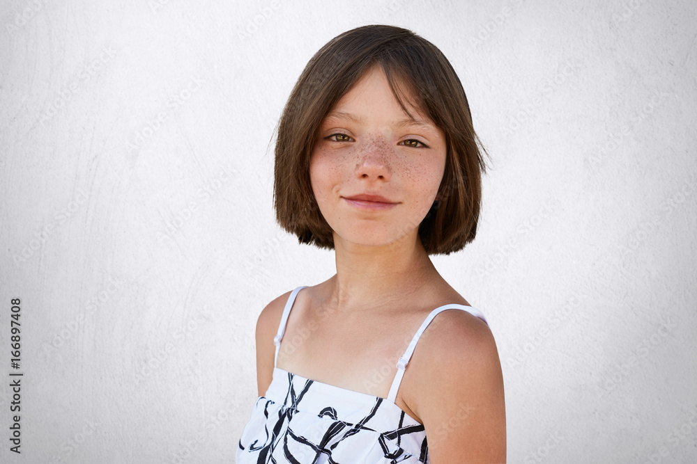 Portrait of freckled little girl with dark short hair, hazel eyes and thin  lips wearing black and white dress, posing at camera against white  background. Adorable girl with freckles. Childhood concept Stock