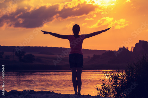 Silhouette of a young woman with arms raised in the backdrop of the setting sun © bravissimos