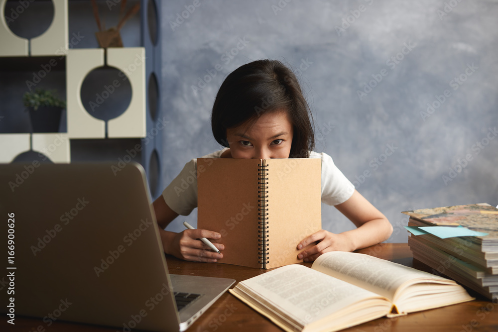Naklejka premium Charming Chinese student girl studying in modern library, working on research project for course paper, sitting at desk with laptop, open book, covering her mouth with copybook and looking at camera