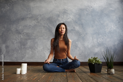 People, meditation and relaxation. Attractive brunette Korean girl wearing top and jeans sitting on wooden floor in half lotus pose, surrounded with candles and plant-pots, meditating with eyes closed