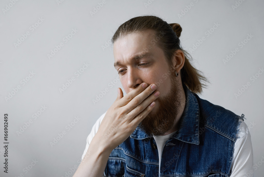 Studio shot of attractive fashionable guy with thick beard and hair knot dressed in denim vest over white t-shirt covering mouth while yawning, feeling bored, sleepy or tired. Body language Studio shot of attractive fashionable guy with thick beard and hair knot dressed in denim vest over white t-shirt covering mouth while yawning, feeling bored, sleepy or tired. Body language