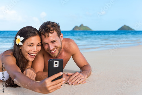 Beach vacation couple taking fun phone selfie on Hawaii vacation. Asian girl Caucasian man relaxing on Lanikai beach, Oahu, on summer holidays using smartphone for pictures together.