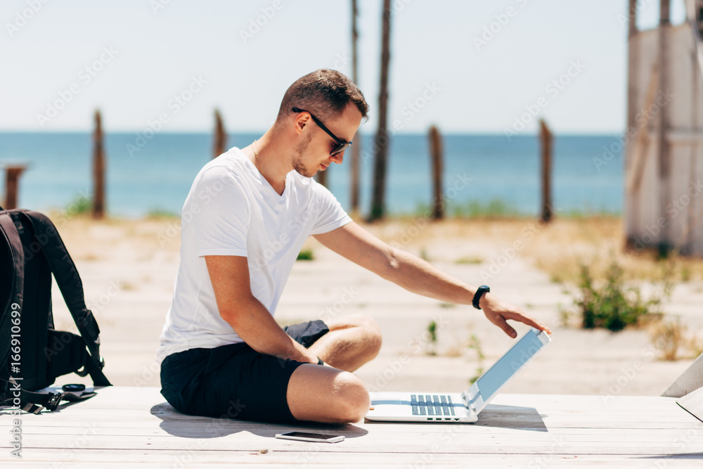 Young man in white T-shirt sits on the beach and working with laptop Young man in white T-shirt sits on the beach and working with laptop