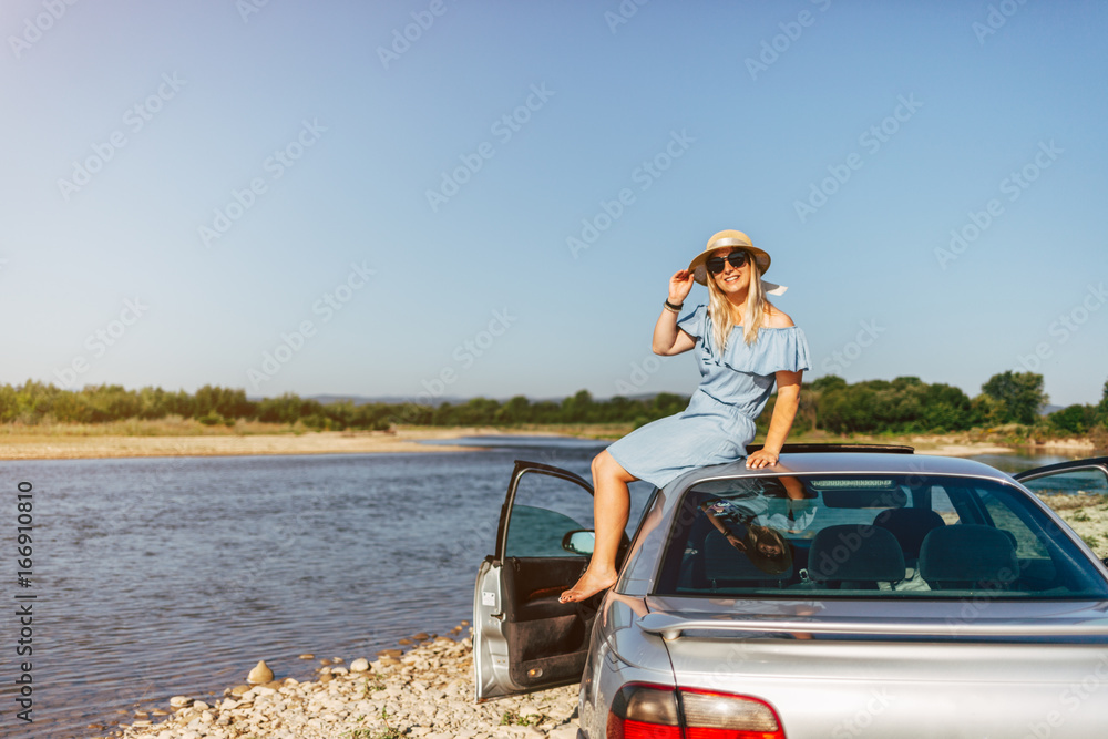 Young traveling woman in a blue dress is sitting by the car by the river Young traveling woman in a blue dress is sitting by the car by the river