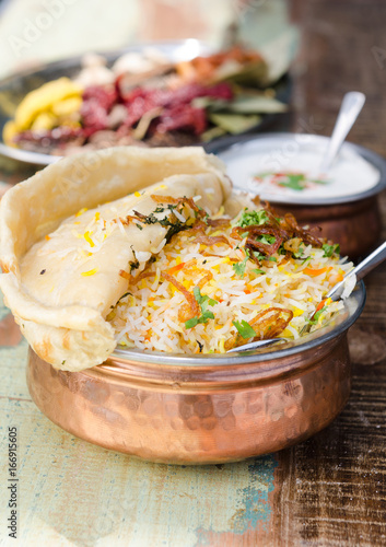 Authentic chicken biryani served with naan bread, fragrant pilau rice and yoghurt, in a metal pot.