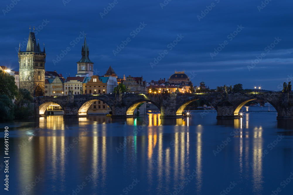 Lit Charles Bridge (Karluv most) and old buildings at the Old Town and their reflections on the Vltava River in Prague, Czech Republic, at dusk.