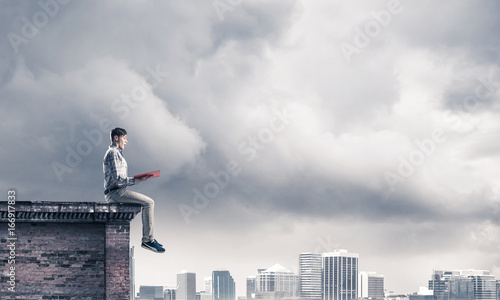 Man on roof edge reading book and cityscape at background