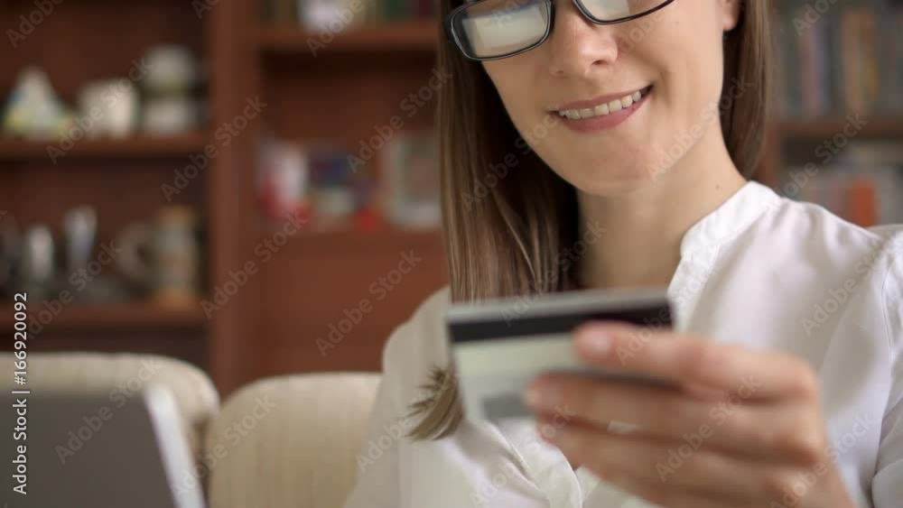 Smiling businesswoman shopaholic in white blouse and glasses sitting on sofa in living room buying online with credit card on laptop. Consumerism internet online purchases Smiling businesswoman shopaholic in white blouse and glasses sitting on sofa in living room buying online with credit card on laptop. Consumerism internet online purchases