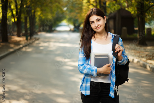 Young cute smiling woman with backpack and notebooks standing in park