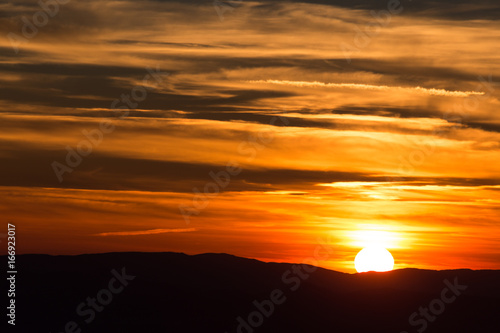 Beautiful sunset with part of the sun coming down behind some mountains and red and orange tones in the sky and clouds