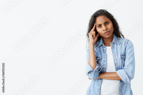 Pretty brunette Afro-american woman with long wavy hair frowning her eyebrows while looking at the camera with placid and thoughtful look. Pensive dark-skinned female with puzzled expression thinking