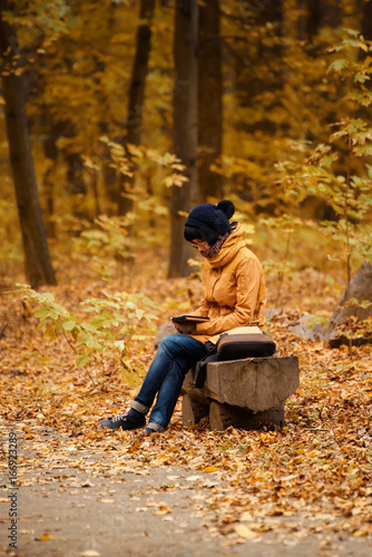 Young stylish hipster girl sitting on the bench in the autumn park and reading electronic book