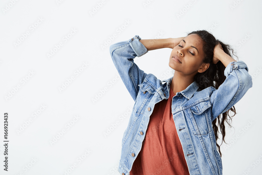 Studio shot of beautiful Afro-american girl in denim jacket with closed eyes playing with her long dark wavy hair. Young woman posing on white background. Studio shot of beautiful Afro-american girl in denim jacket with closed eyes playing with her long dark wavy hair. Young woman posing on white background.
