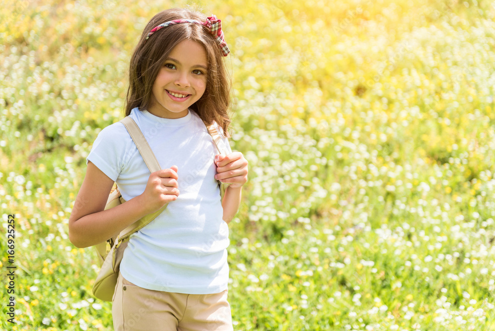Joyful female kid walking on grassland with rucksack Joyful female kid walking on grassland with rucksack