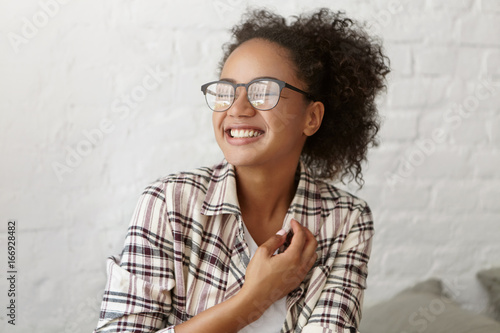 Joyful mixed race woman with curly pony tail wearing casual checkered shirt smiling pleasantly and closing her eyes with excitement, feeling relaxation and joy while being glad to meet with friends