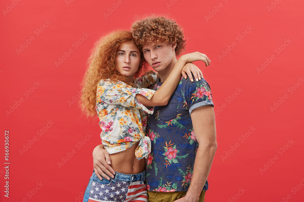 Stlyle and fashion concept. Indoor studio shot of fashionable young red-haired European man and woman wearing trendy summer clothing, posing at red wall, embracing each other and looking at camera