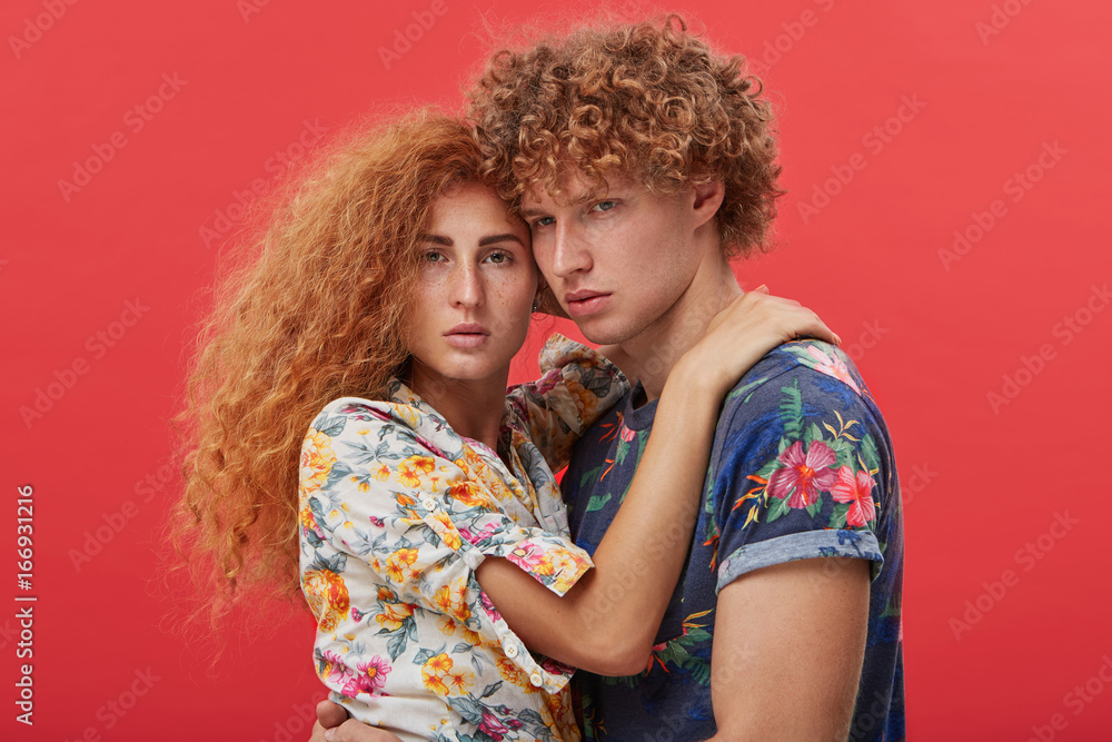 Headshot of two young people in love modeling, posing together during photo shooting in studio: serious redhead male in floral t-shirt standing close to pretty girl who holding hands around his neck Headshot of two young people in love modeling, posing together during photo shooting in studio: serious redhead male in floral t-shirt standing close to pretty girl who holding hands around his neck