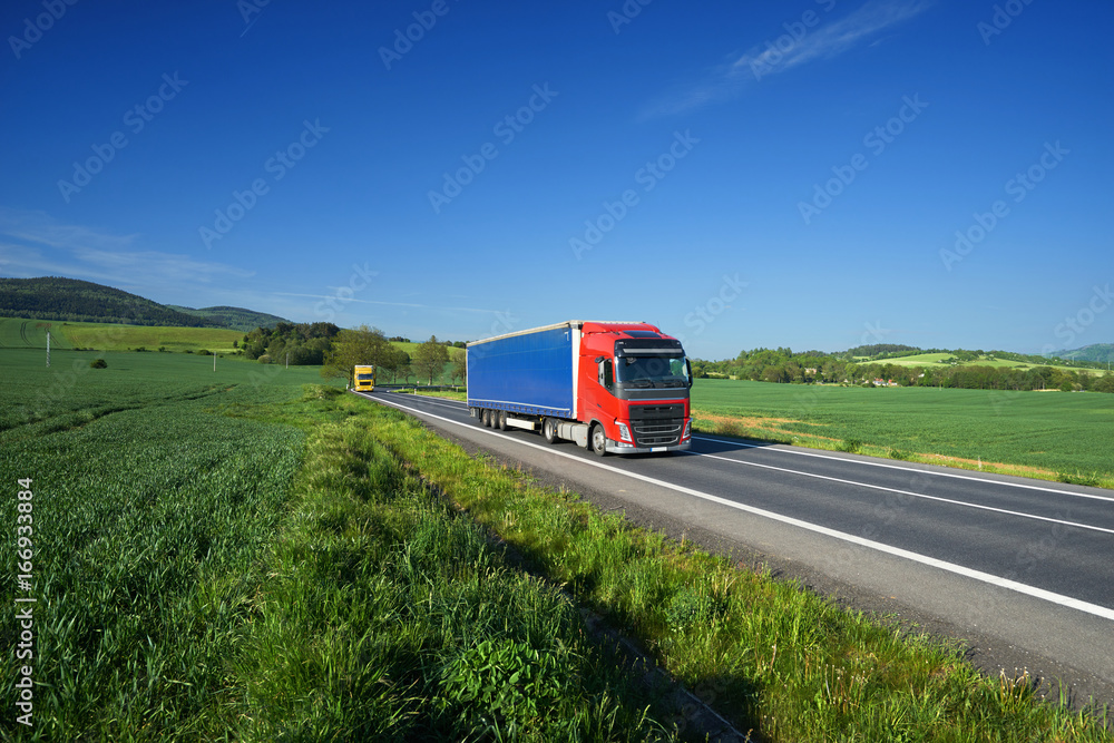 Trucks transporting goods on the asphalt road between green fields in a rural landscape under a blue sky