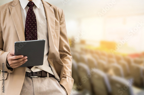 businessman using tablet or mobile phone, blurred of conference hall or seminar room with attendee background.