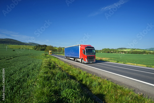 Obraz na płótnie Trucks transporting goods on the asphalt road between green fields in a rural landscape under a blue sky w salonie