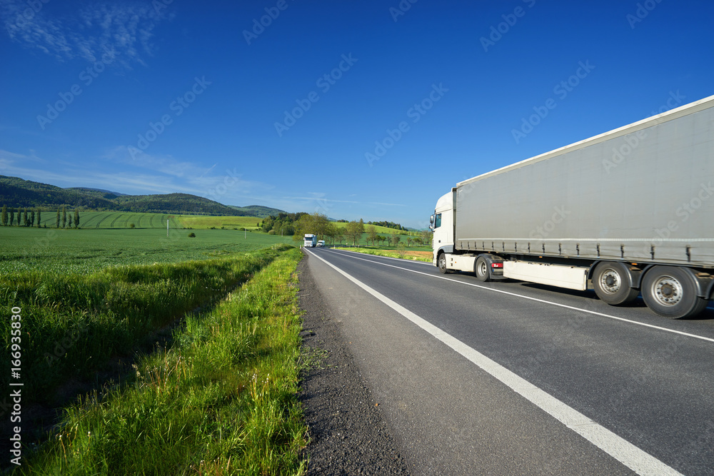 White truck transporting goods on the asphalt road between green fields in rural landscape with wooded mountains in background