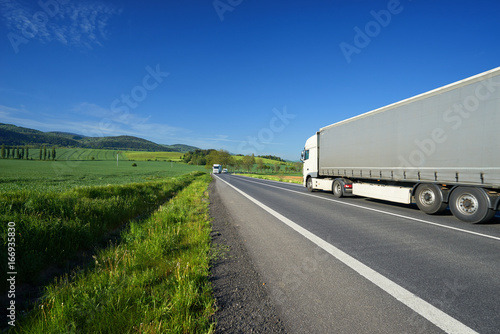 White truck transporting goods on the asphalt road between green fields in rural landscape with wooded mountains in background