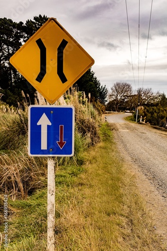 Title: One lane/right of way road signs. Top, yellow with road narrowing symbol. Bottom, blue sign with white and red arrows. Cloudy sky, trees, gravel road in background