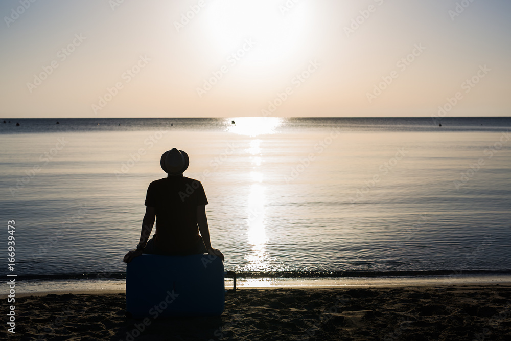 Back view silhouette of man sitting on suitcase on blue sea outdoors background. Arriving at destination