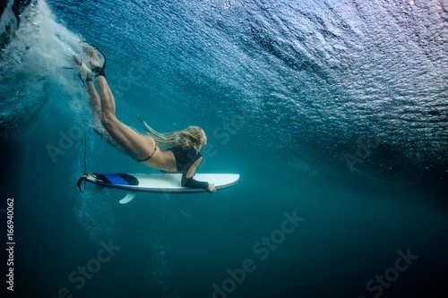 Blonde girl Surfer holding white surf board Diving Duckdive under Big Beautiful Ocean Wave. Turbulent tube with air bubbles and tracks after sea wave crashing. photo