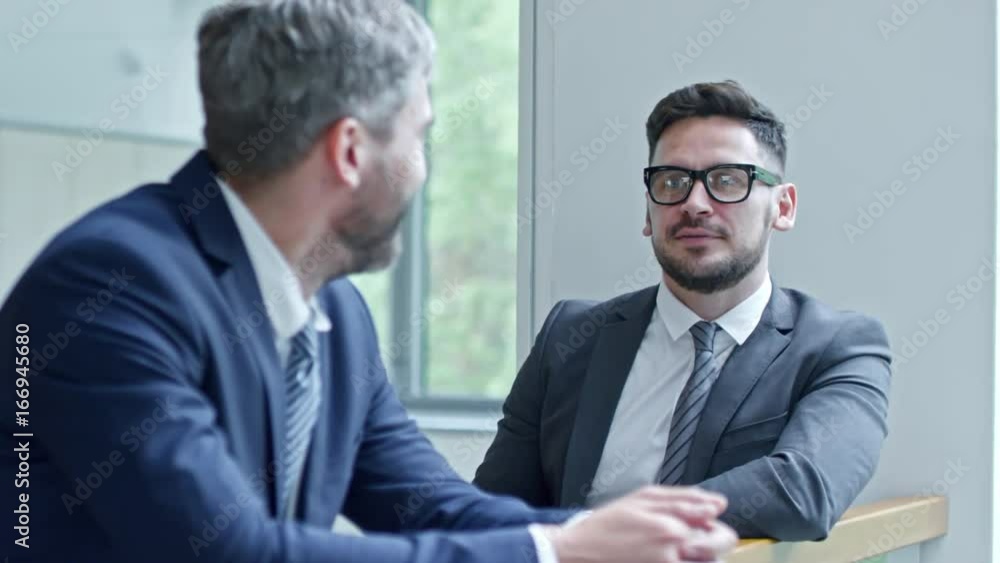 PAN of friendly businessman in glasses and suit and his male colleague in professional clothing leaning on railing and chatting PAN of friendly businessman in glasses and suit and his male colleague in professional clothing leaning on railing and chatting