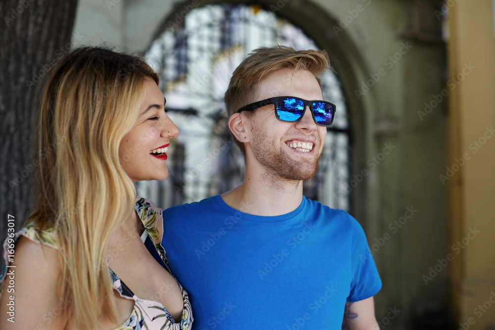 People, love, relationships, fun, relaxation and happiness. Pretty girl in flower design top laughing, standing in urban setting close to her handsome boyfriend with stubble wearing stylish shades People, love, relationships, fun, relaxation and happiness. Pretty girl in flower design top laughing, standing in urban setting close to her handsome boyfriend with stubble wearing stylish shades